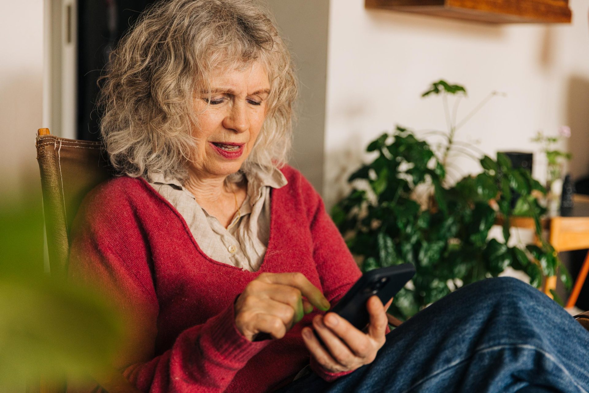 Eldre kvinne med krøllete grått hår ved hjelp av en smarttelefon i en koselig stue med planter i bakgrunnen.