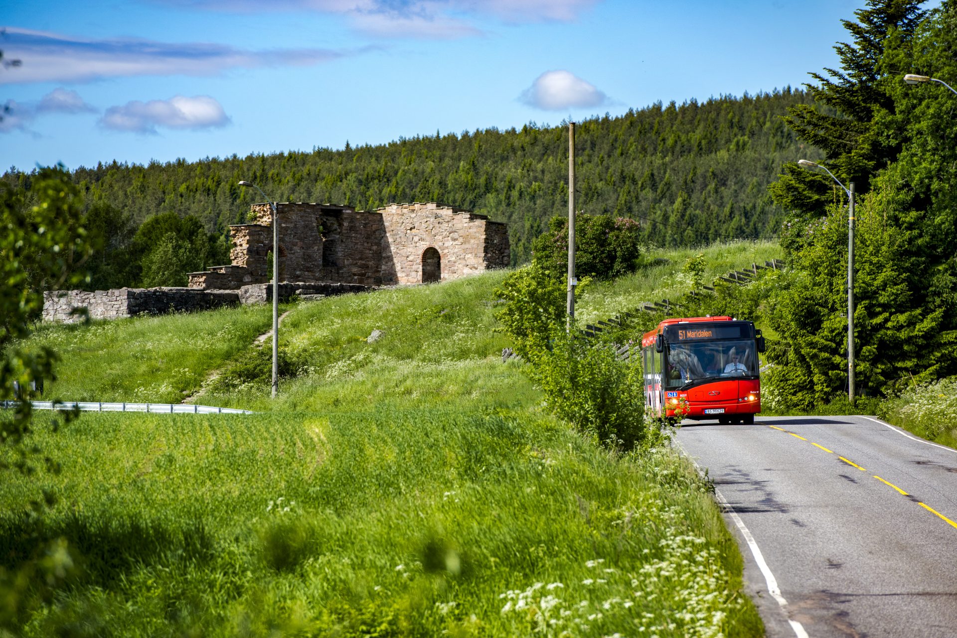 En rød buss kjører på en vei forbi gamle steinruiner i et frodig grønt landskap under en klarblå himmel.