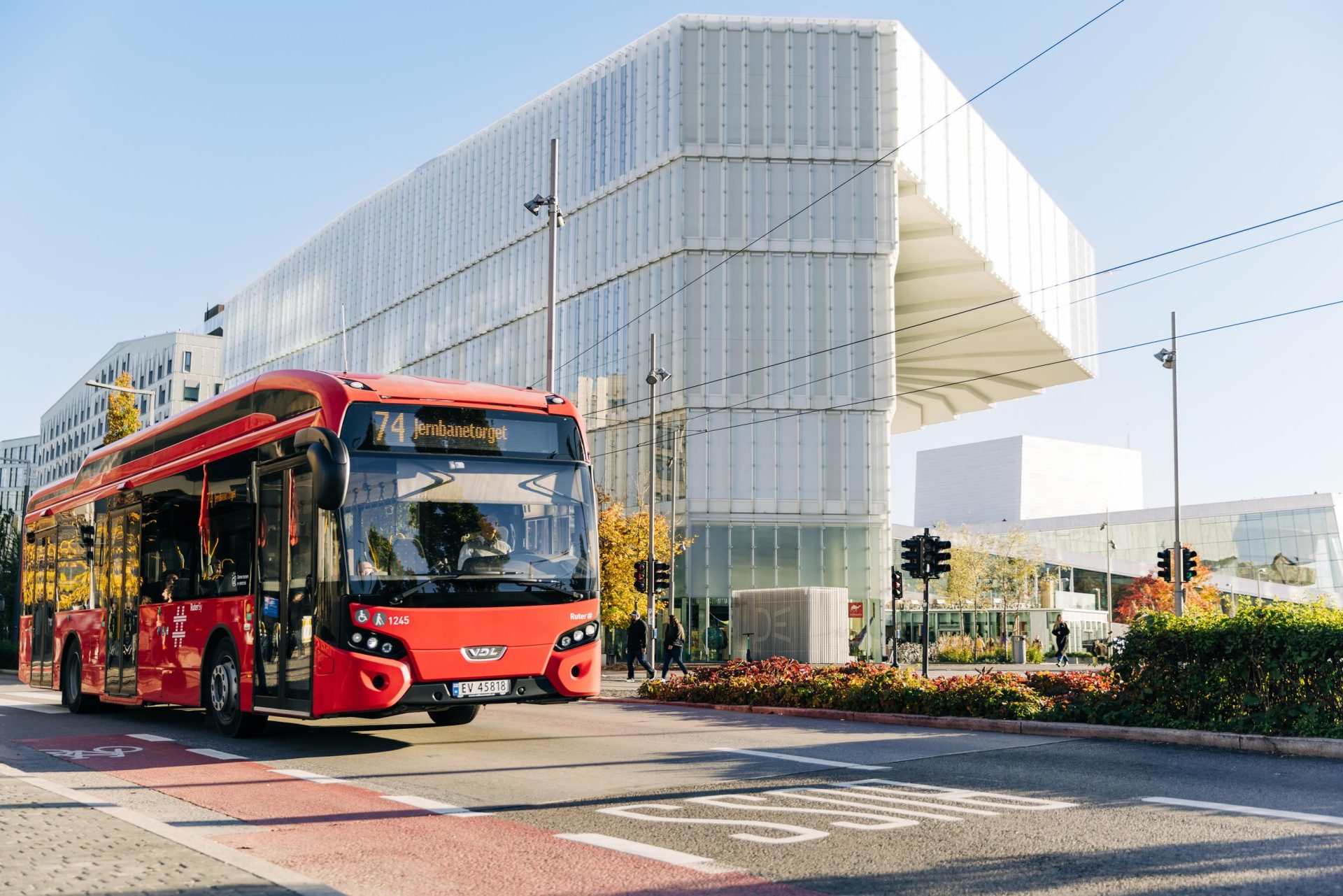 En rød buss merket med nummer 74 kjører gjennom en bygate nær en moderne hvit arkitekturbygning på en solrik dag.