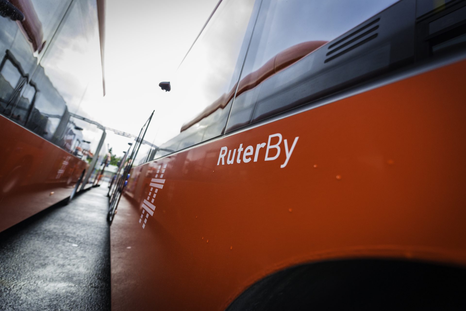 Oransje bybuss med logoen «ruterby» synlig, parkert på en gate under overskyet himmel, reflektert på overflaten til en annen buss.