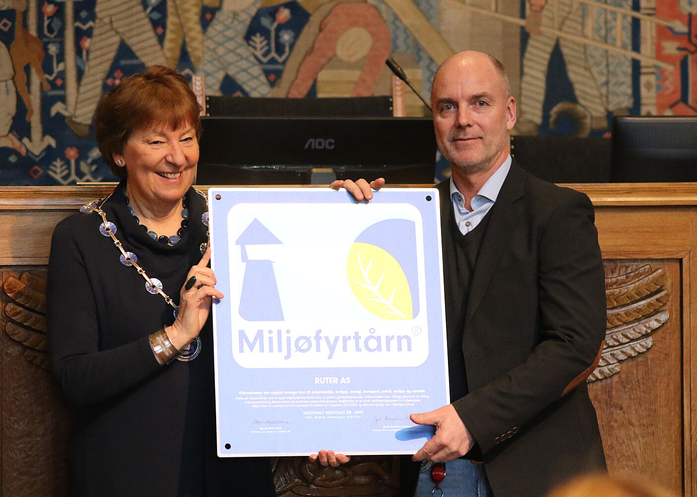 En kvinne og en mann som holder et stort sertifikat med et miljøvennlig symbol, står i et rom med utsmykkede veggtepper.