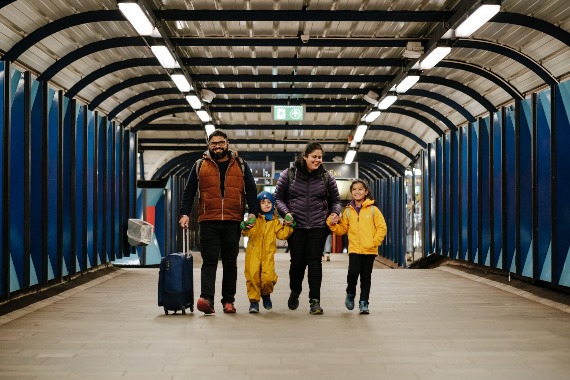 En familie på fire går gjennom en T-banestasjonstunnel, holder hender og bærer en koffert.