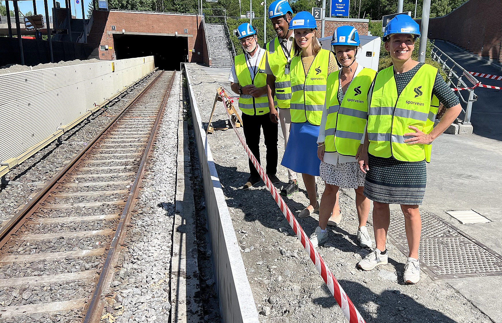 Fem bygningsarbeidere i hjelme og refleksvester står ved et jernbanespor og smiler til kamera.