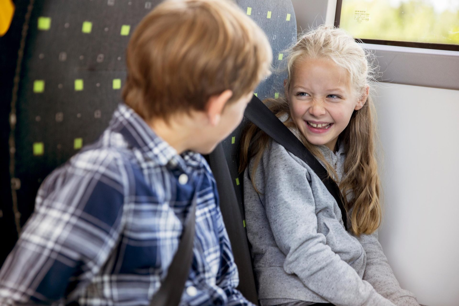 En ung gutt og jente som smiler og snakker på en buss, både sittende og med bilbelte. jenta virker underholdt, og gutten står vendt mot henne.