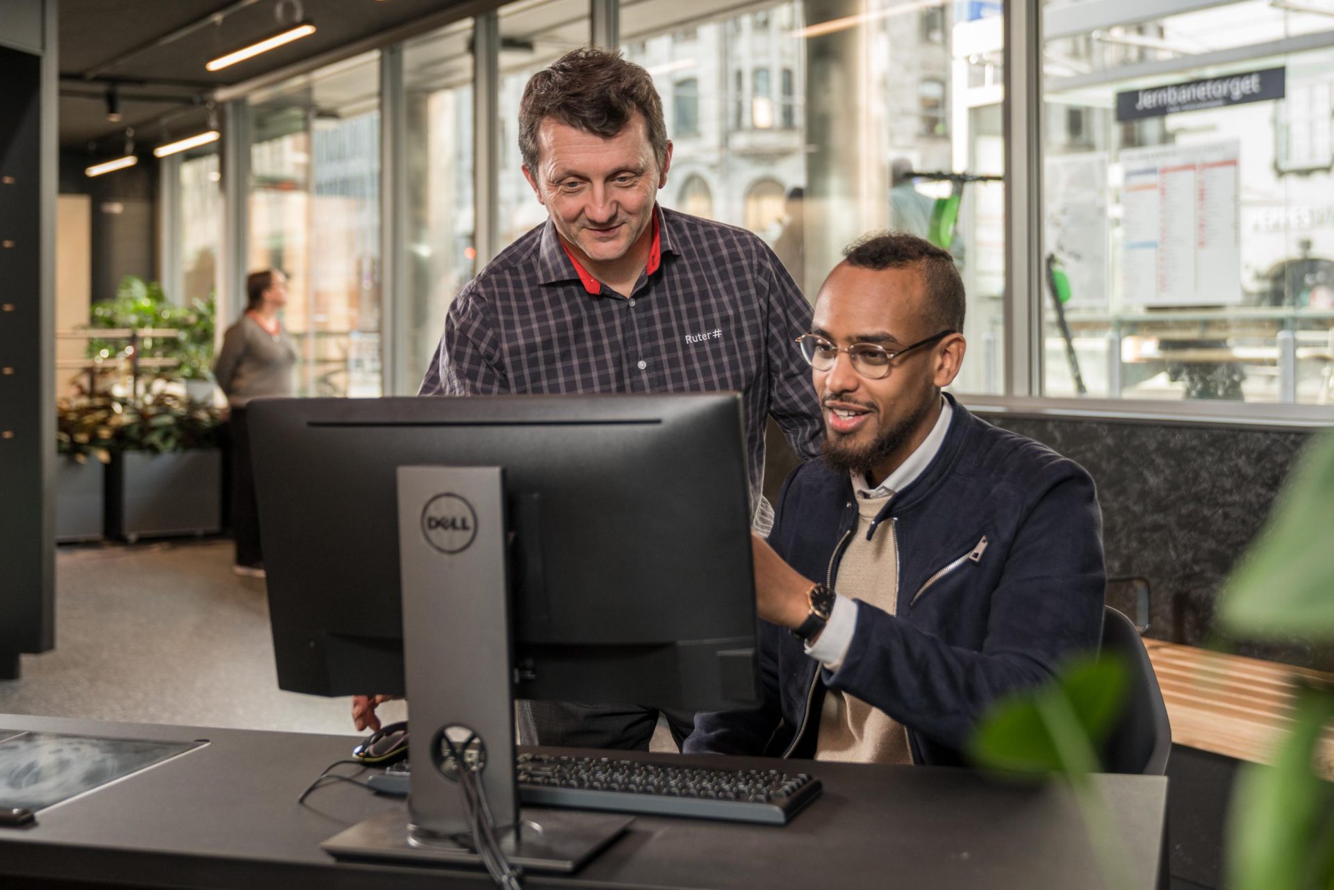 To menn, en eldre og en yngre, smiler mens de ser på en dataskjerm i et moderne kontormiljø.