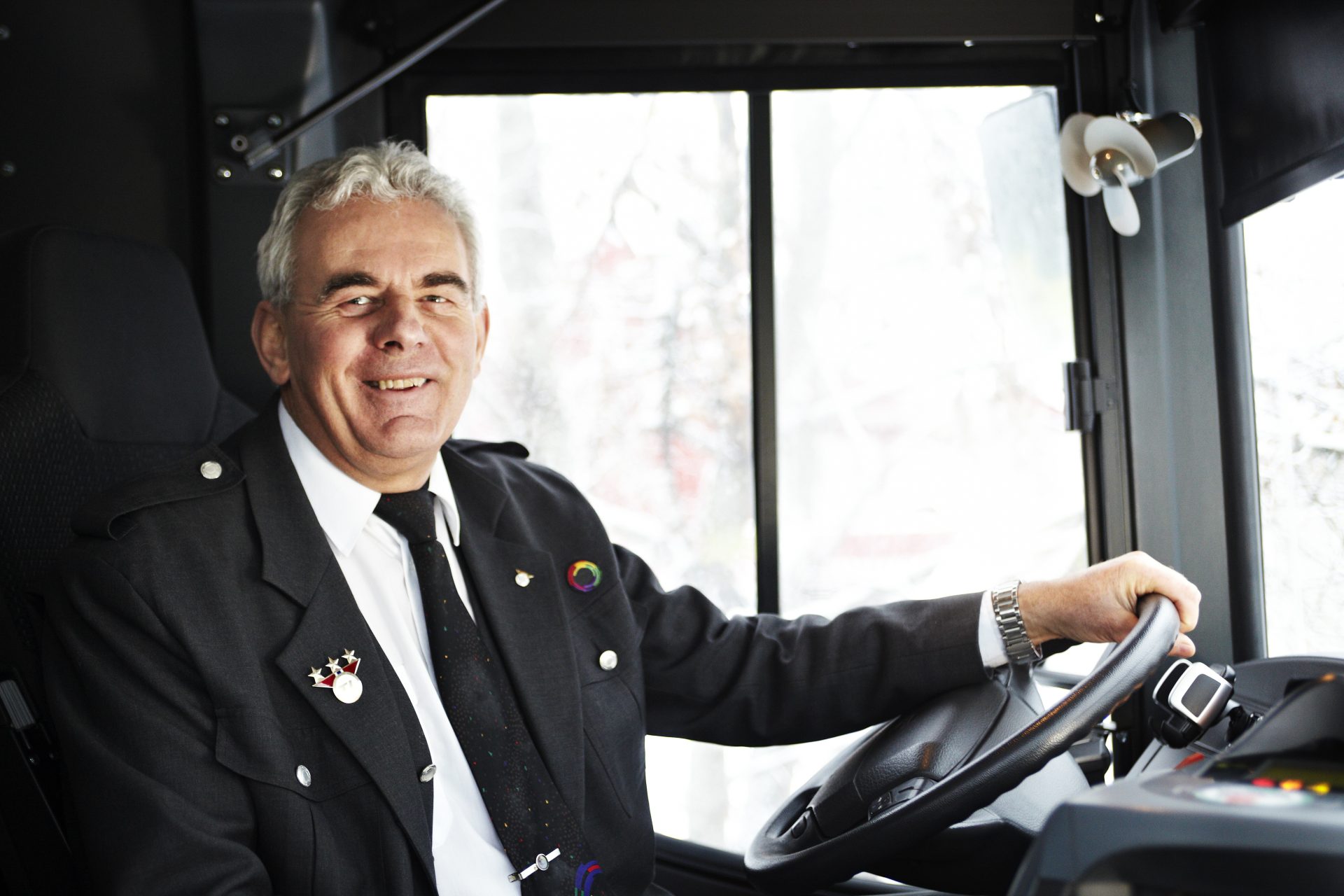 En blid bussjåfør i uniform sitter ved rattet i en buss og ser på kameraet med et smil.