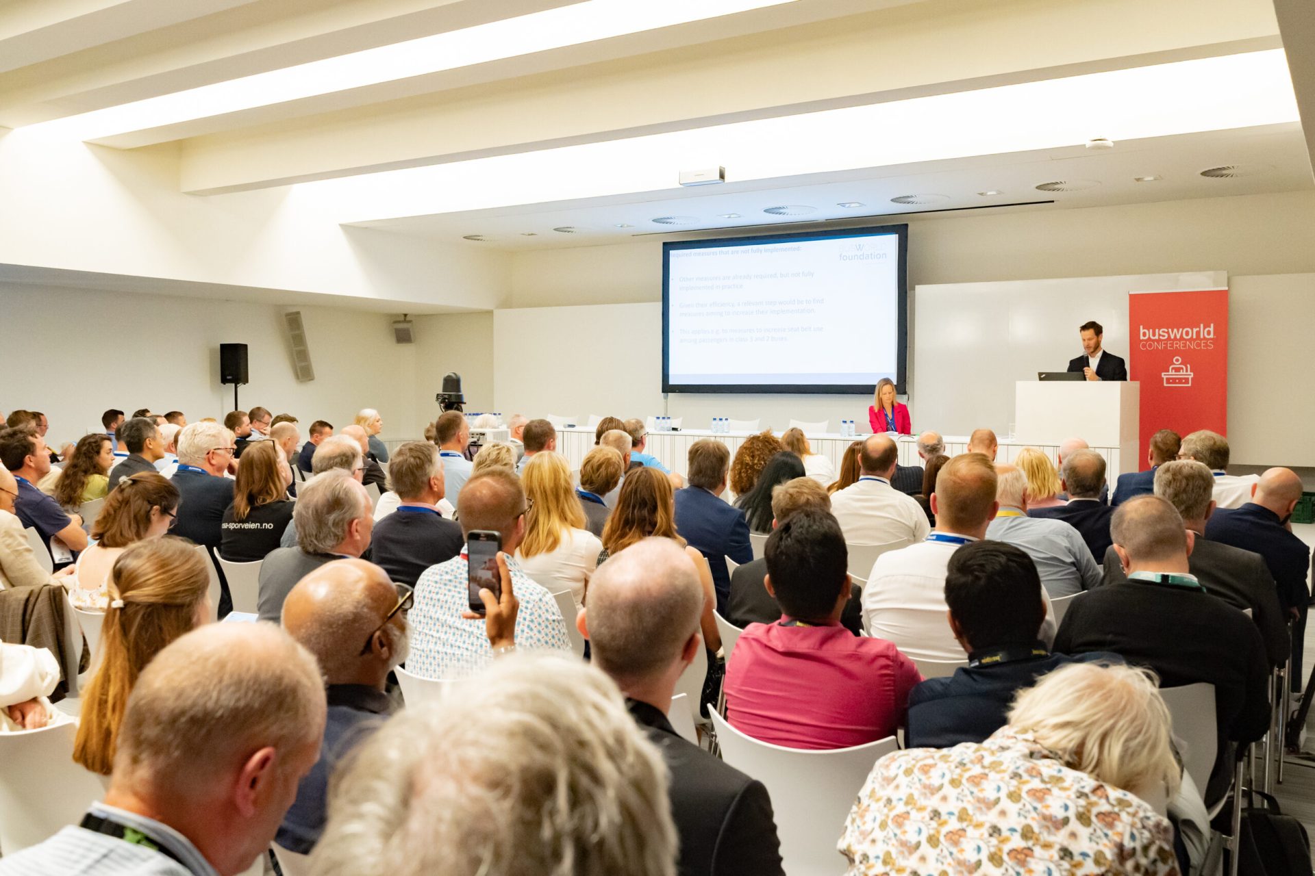 Et konferanserom med deltakere vendt mot en presentatør som taler ved et podium under et profesjonelt arrangement, med en projeksjonsskjerm som viser en presentasjon.