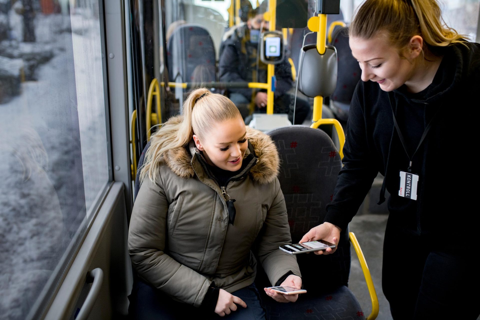 Billettkontrollør sjekker en kvinnelig passasjers billett på en buss, med andre reisende synlige i bakgrunnen.