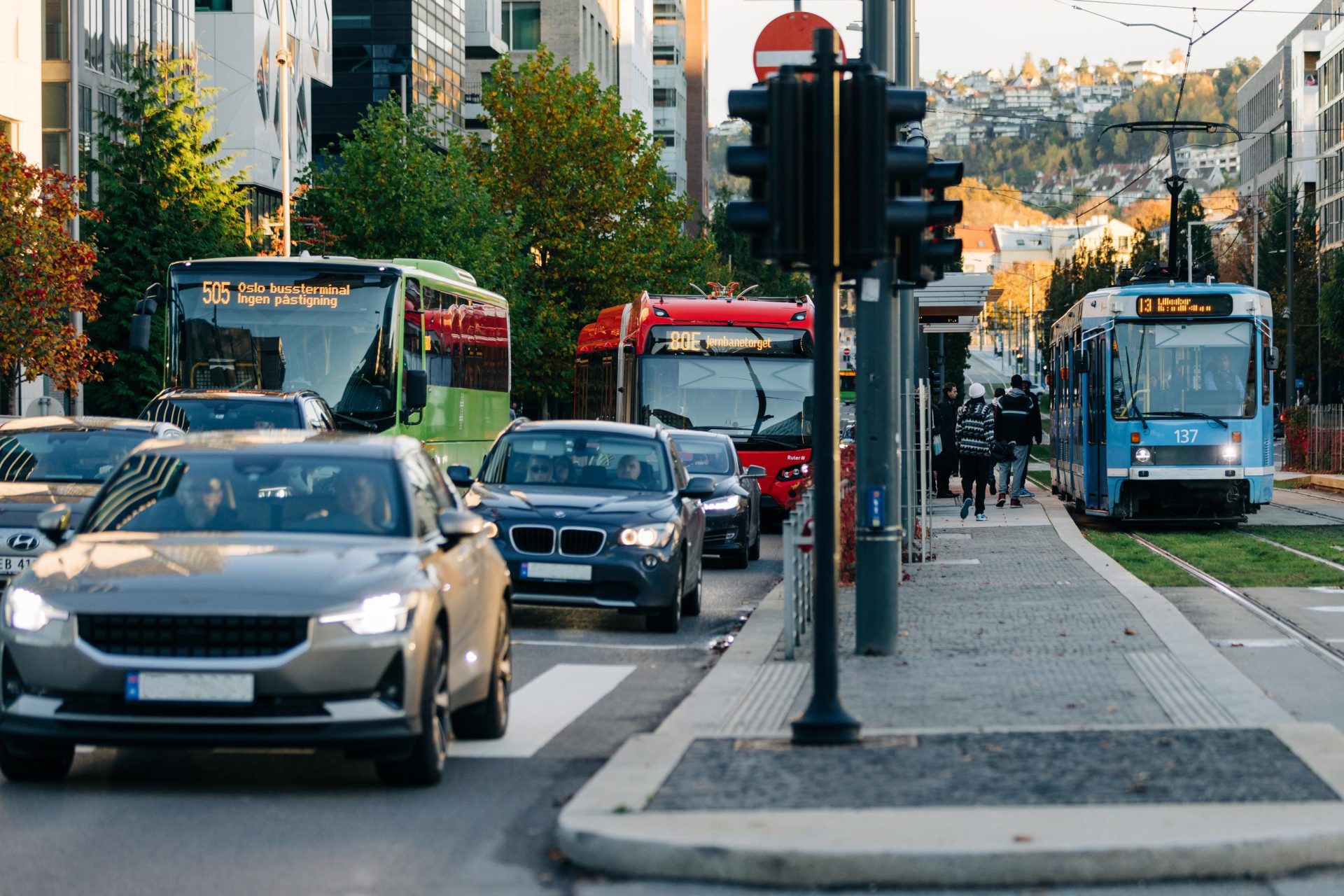 Trafikkscene i en by med biler, busser og en trikk som beveger seg langs en gate, med fotgjengere som venter på å krysse i et lyskryss.