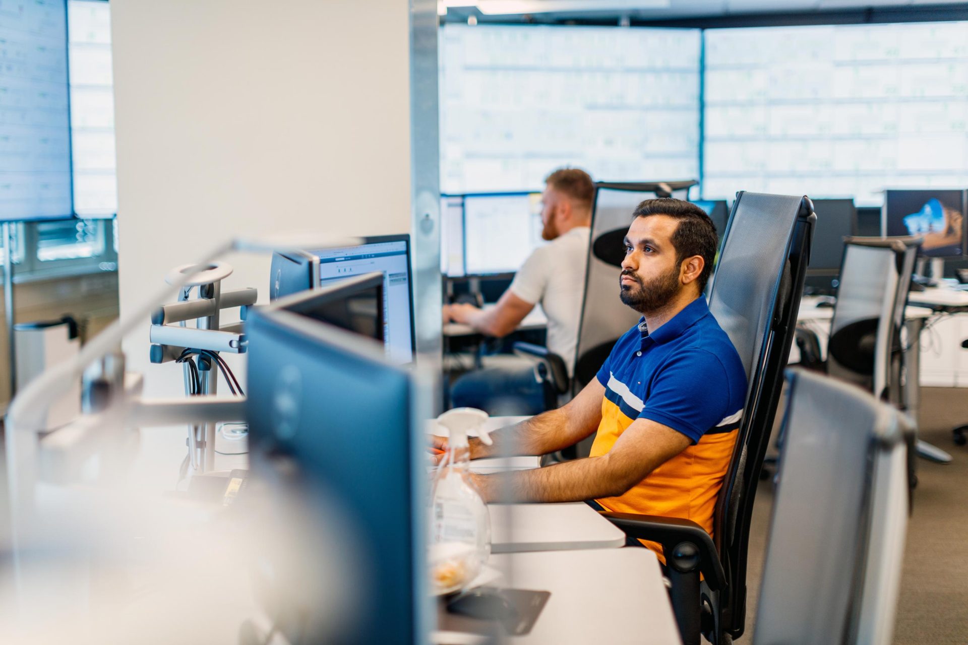 A man in a blue and orange shirt works at a computer in a modern office with other colleagues around him.