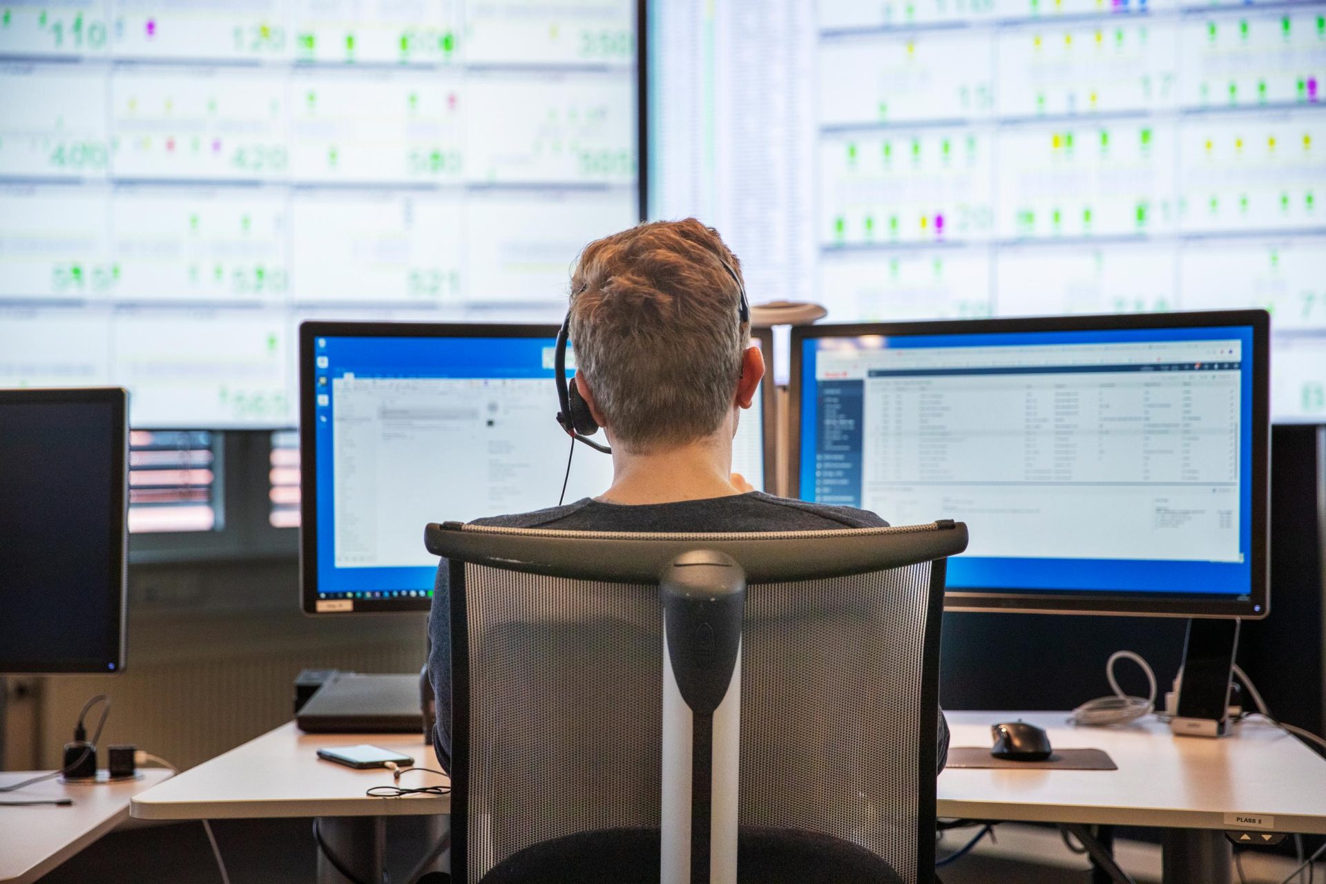 Man wearing headphones sitting at a desk, facing several computer monitors showing data and charts in an office environment.