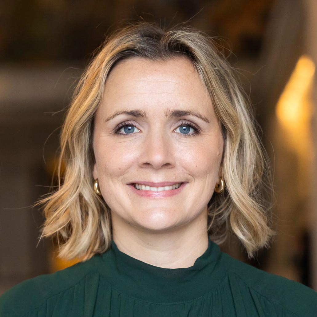 Portrait of a smiling woman with shoulder-length blonde hair, wearing a green blouse and gold earrings, indoors with a blurred background.