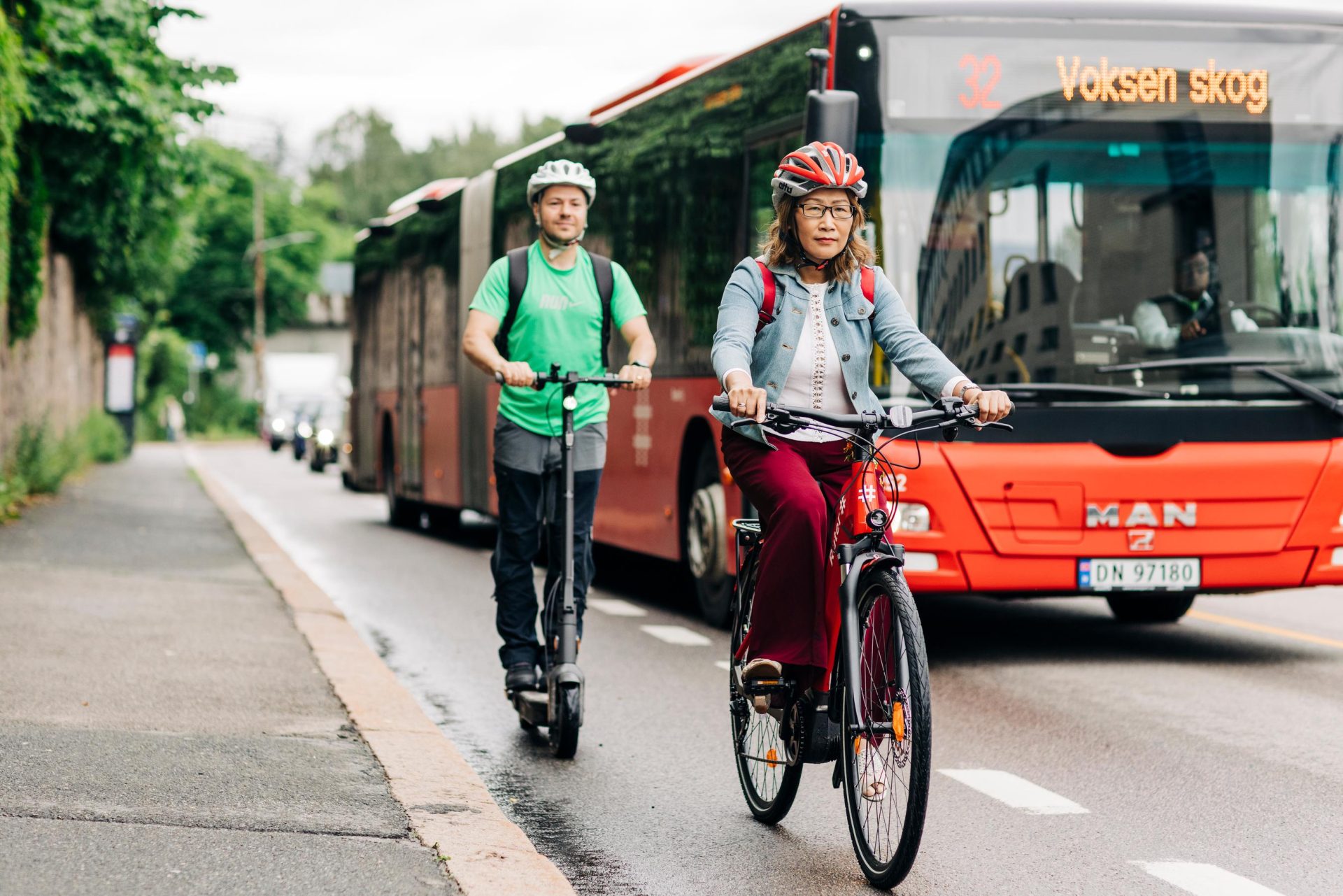 En mann på sparkesykkel bak en kvinne på en sykkel i en bygate ved siden av en buss i bevegelse; trær langs veien.