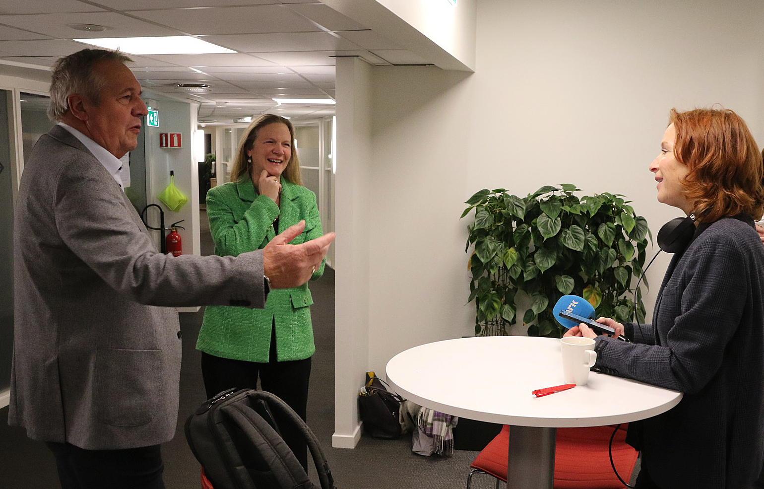 Two women and a man talking in an office corridor, a woman laughing while the man gestures animatedly, a small table with a pen and a cup between them.