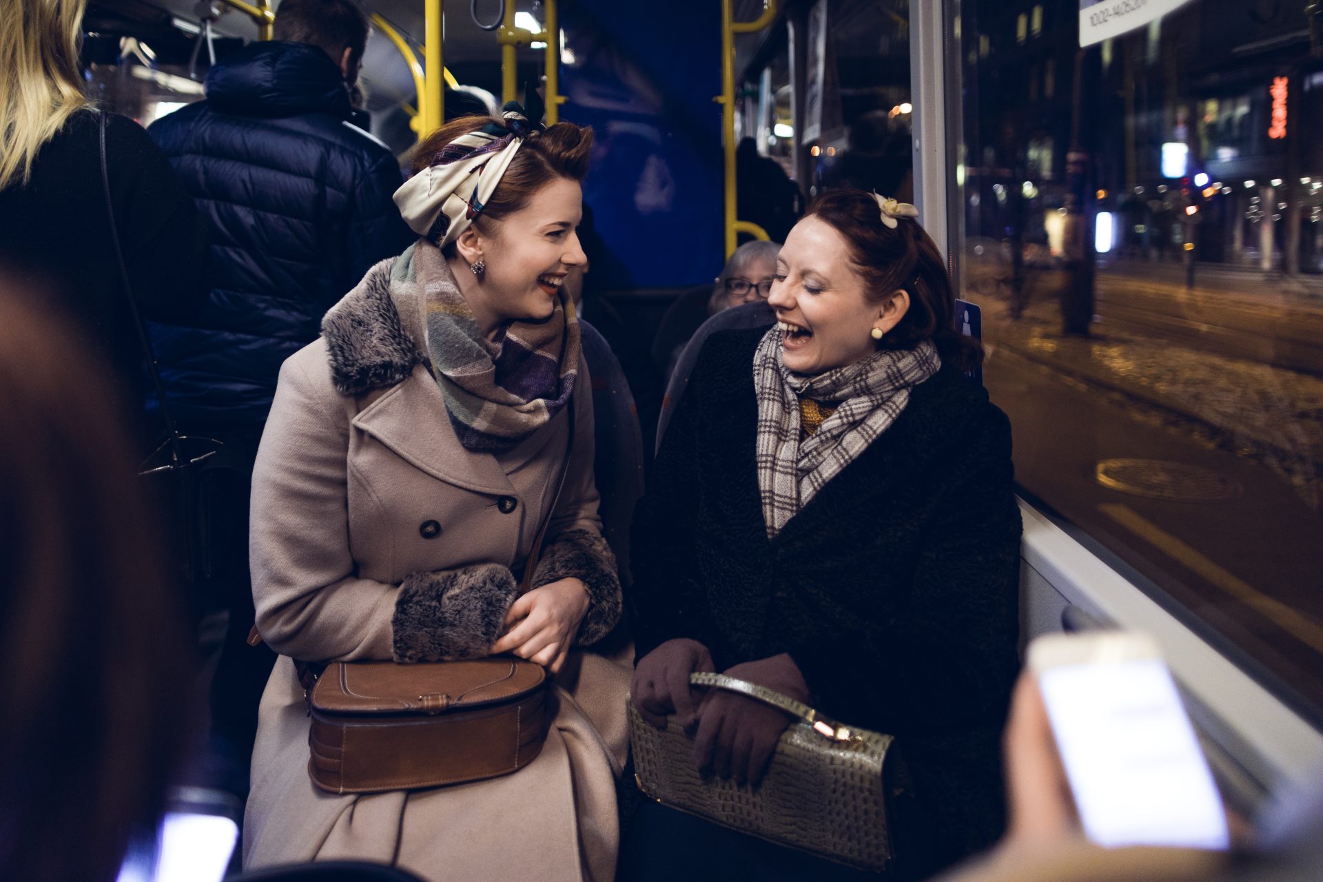 Two women laughing and chatting on a city bus at night, surrounded by other passengers.