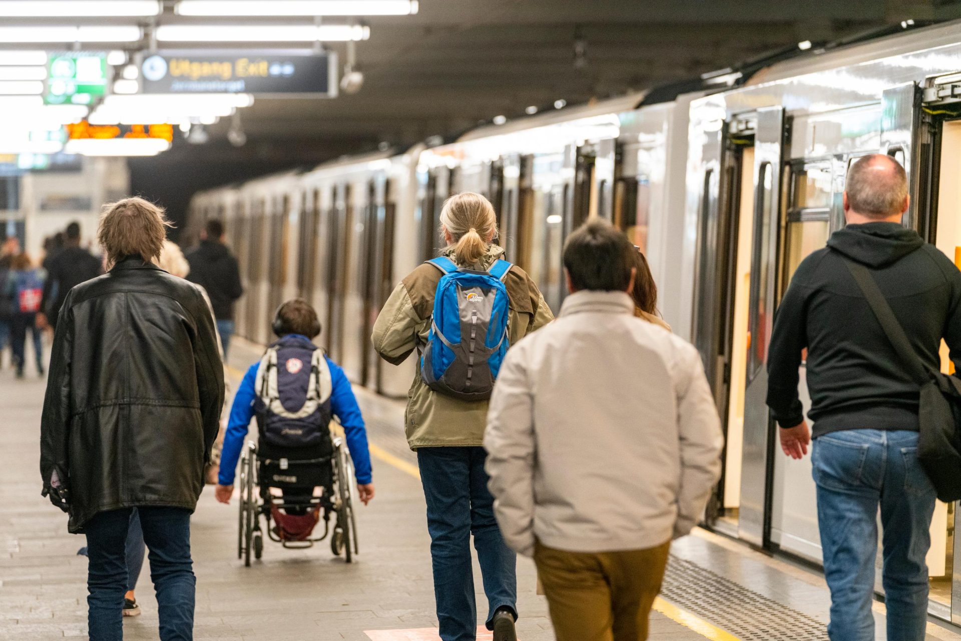 Passasjerer som går på en T-baneplattform ved siden av et stillestående tog, inkludert en person i rullestol og andre med ryggsekker.