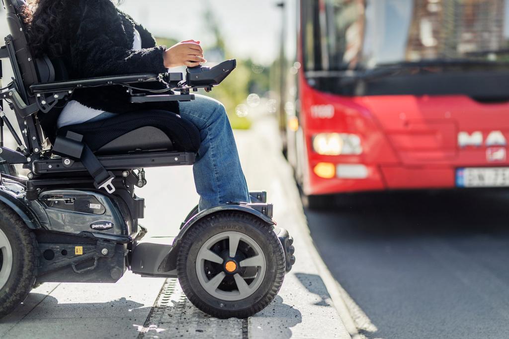 Person i motorisert rullestol venter på et busstopp, med en rød buss synlig i bakgrunnen.