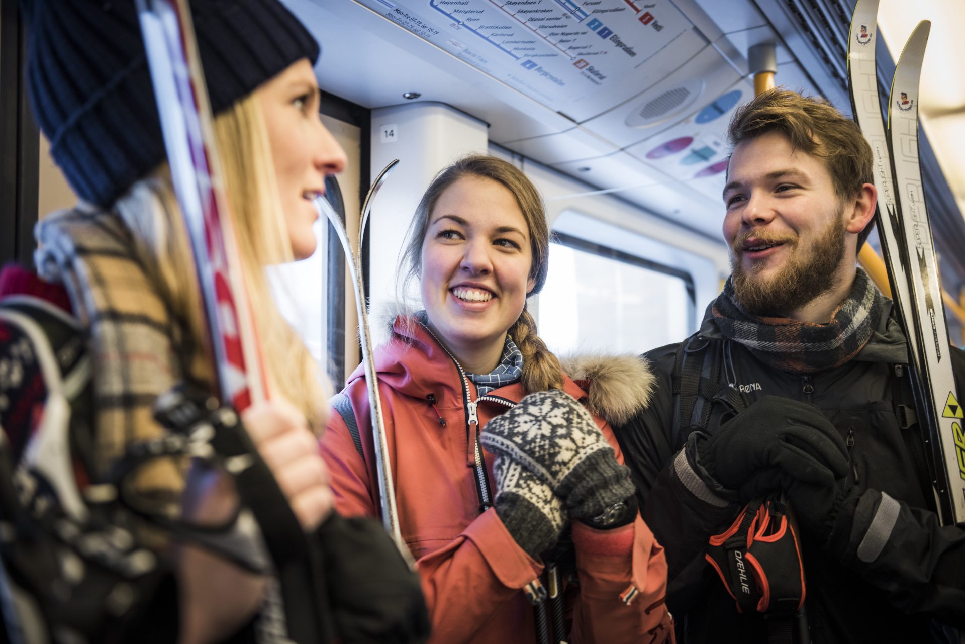 Three young adults chatting happily inside a train, dressed in winter clothes and holding ski equipment.