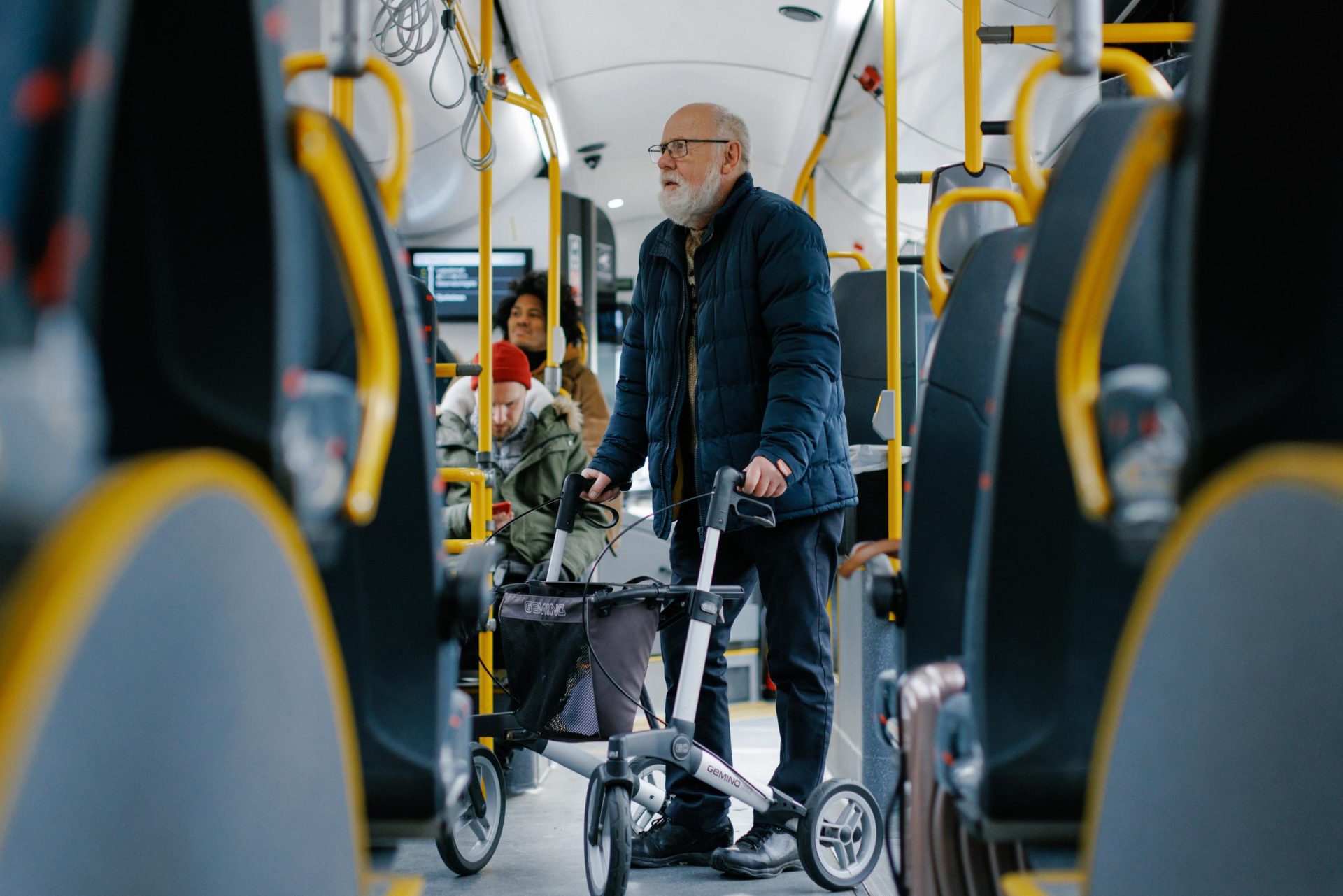 An elderly man with a walker stands in the aisle of a bus, with other passengers around him.