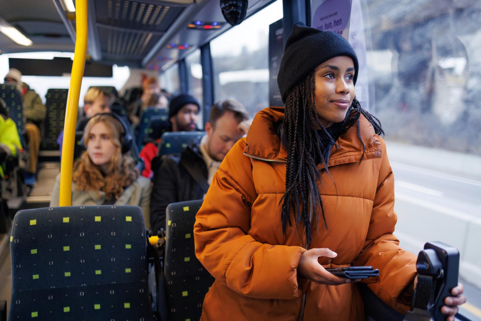 En ung kvinne i en oransje jakke som holder en smarttelefon ser ut av vinduet på en buss fylt med passasjerer.