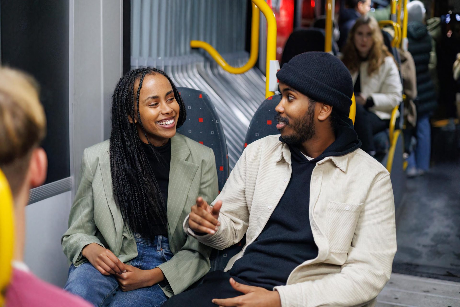 En mann og en kvinne, begge smilende, deltar i samtale på en bybuss. kvinnen har flettet hår og har på seg en grønn jakke; mannen har på seg en beige jakke og en svart lue.