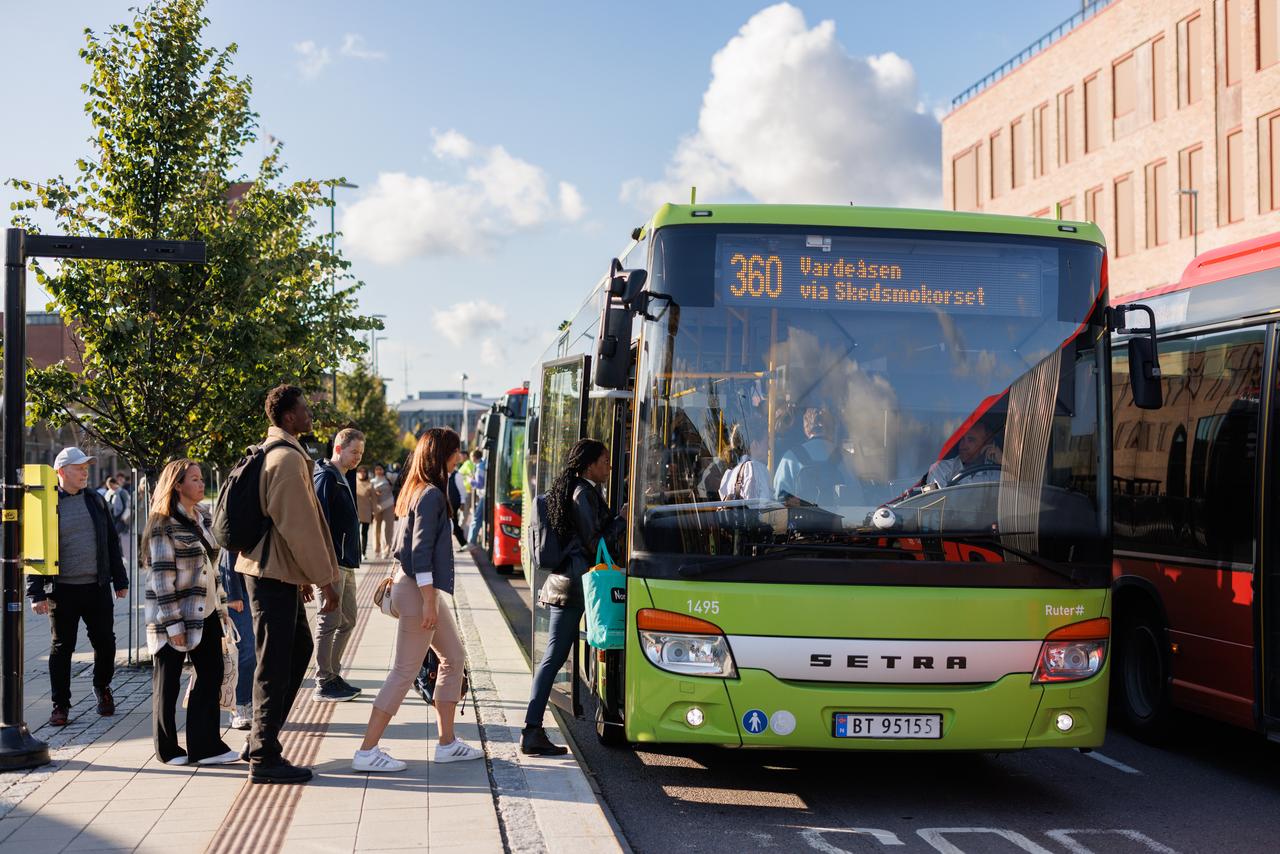 Passasjerer som går ombord på en grønn buss merket rute 360 ved et busstopp på en solrik dag, med en folkemengde som står i kø.