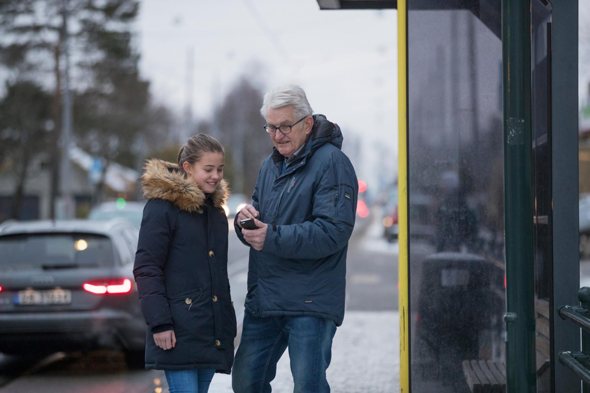 En tenåringsjente og en eldre mann undersøker en smarttelefon sammen på et busstopp, med biler som passerer i bakgrunnen.