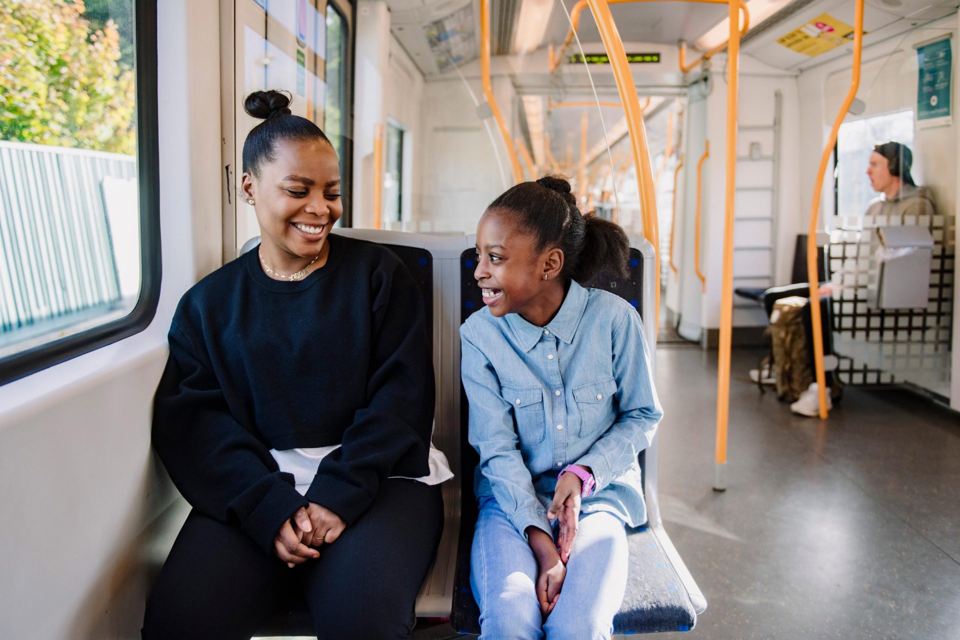 Two women smiling and talking on a train, with other passengers visible in the background.