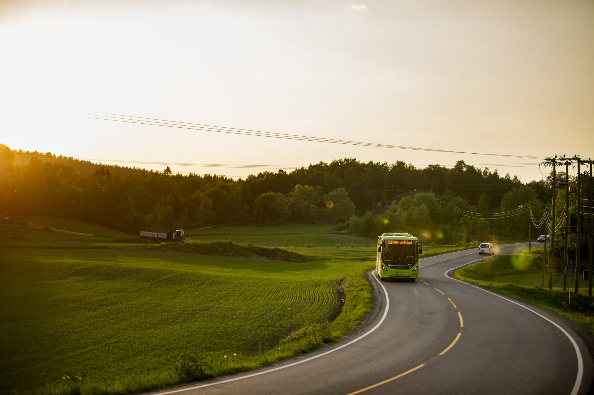 En buss kjører langs en svingete vei gjennom frodig grønt landskap ved solnedgang, med kraftledninger og kjøretøy synlige i bakgrunnen.