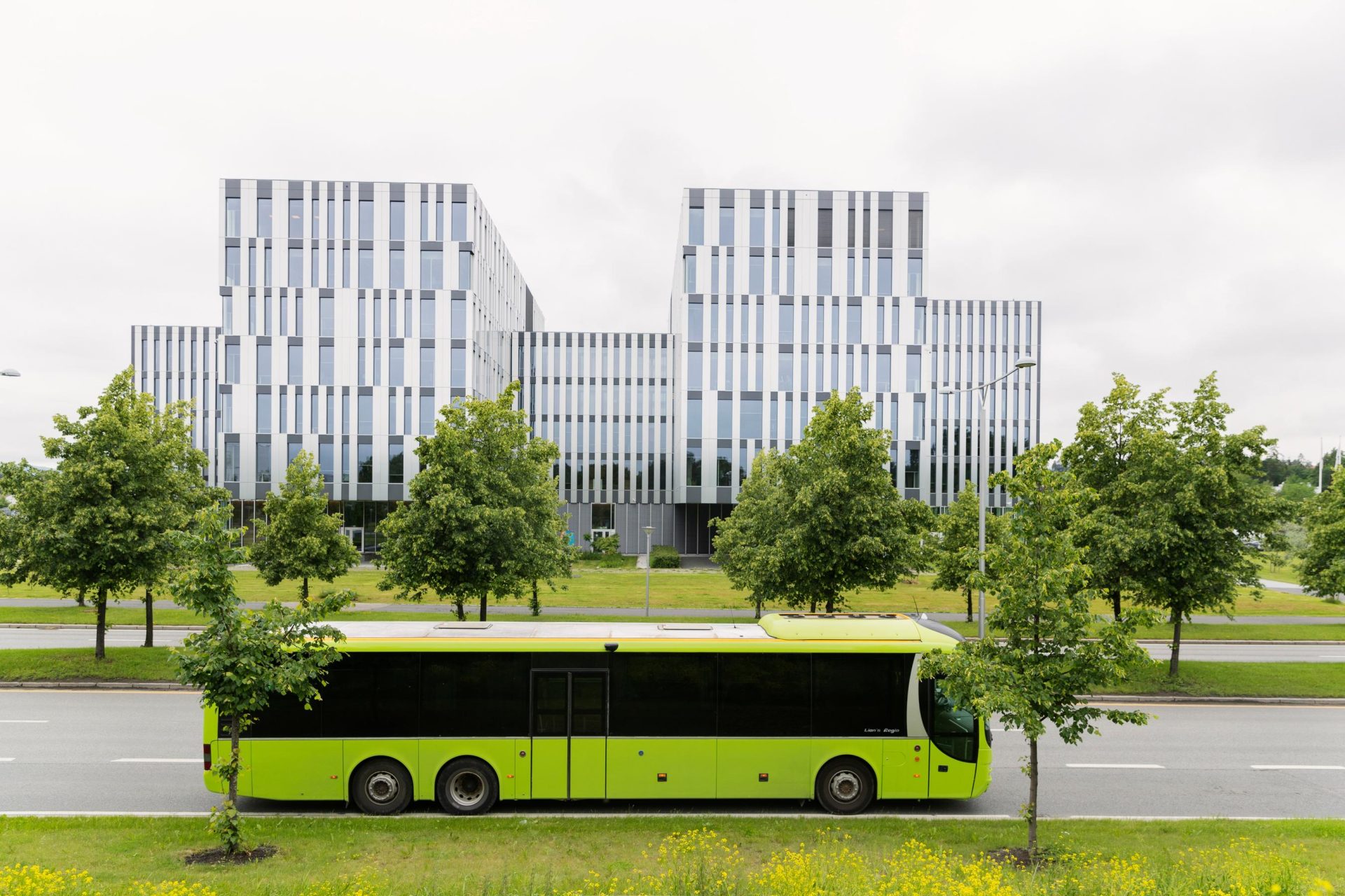 Moderne kontorbygg med symmetrisk design og vertikale aksenter bak en knallgrønn buss på en byvei omkranset av trær.
