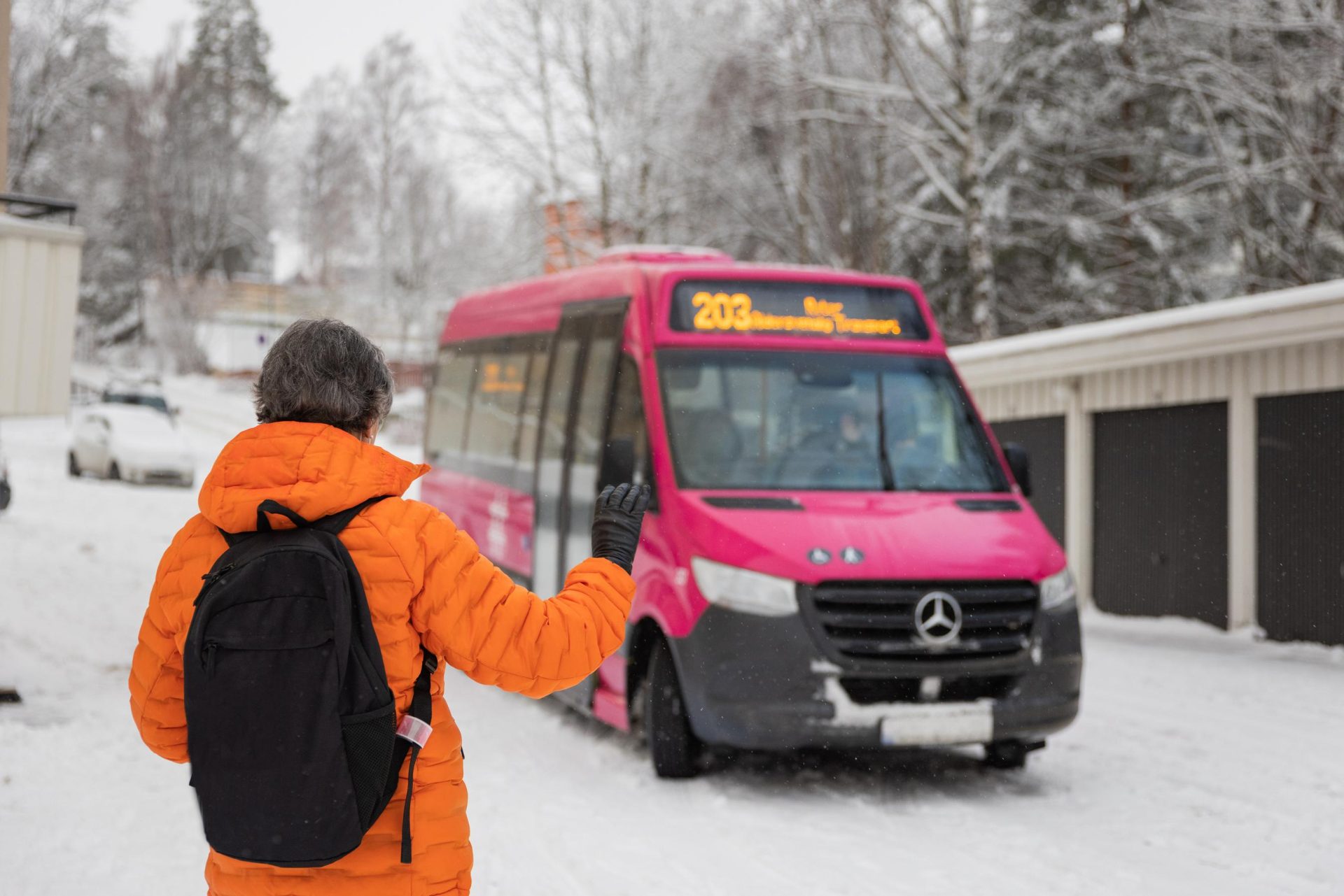 En person i oransje jakke og ryggsekk vinker til en rosa minibuss på en snørik dag.