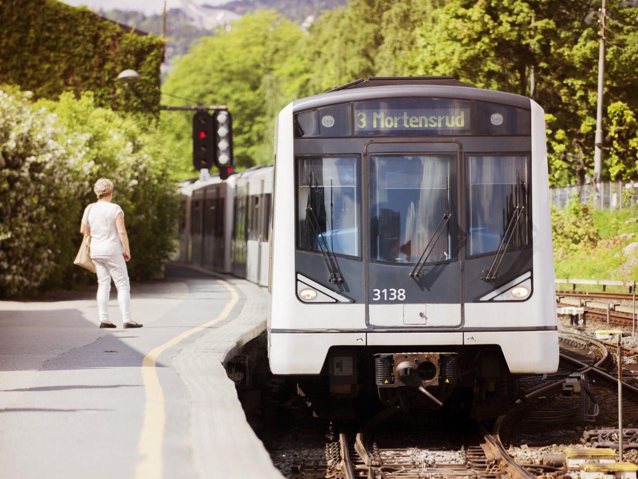 En T-bane merket «3 mortensrud» nærmer seg en stasjon mens en person venter på å gå ombord, med grønt bladverk i bakgrunnen.