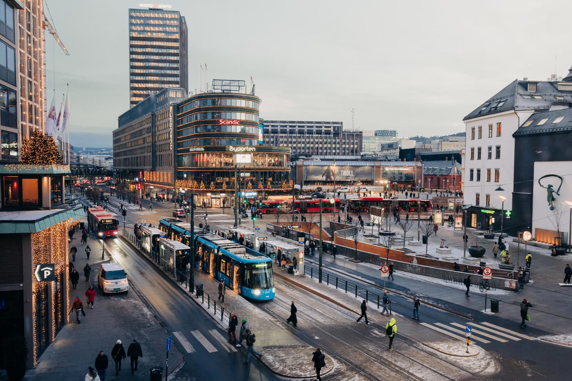 En travel byscene på Jernbanetorget i skumringen med trikker og fotgjengere, med moderne bygninger og gatelys, om vinteren.