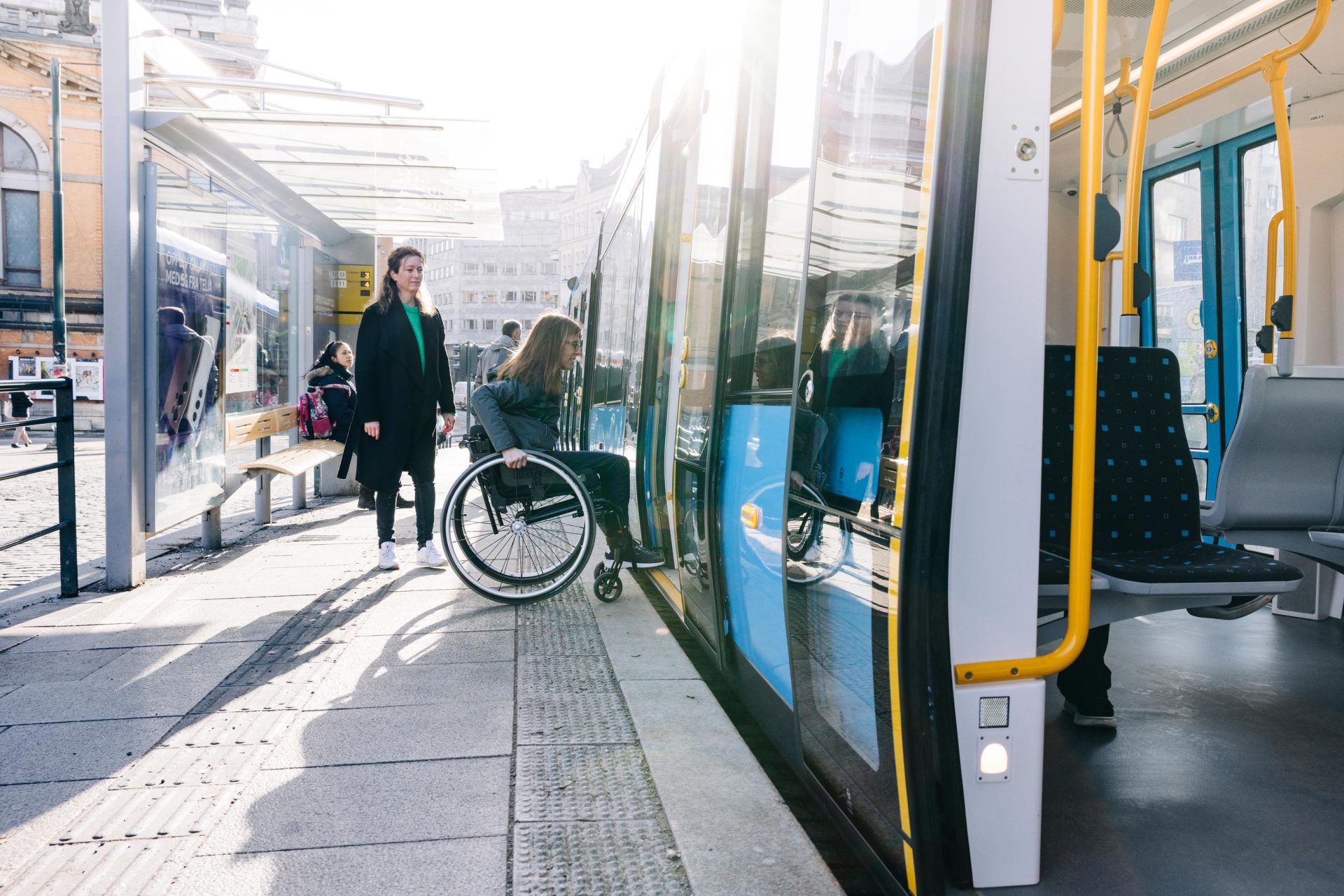 En person i rullestol som går ombord på en moderne trikk, med en annen passasjer som går ved siden av, i solfylte urbane omgivelser.