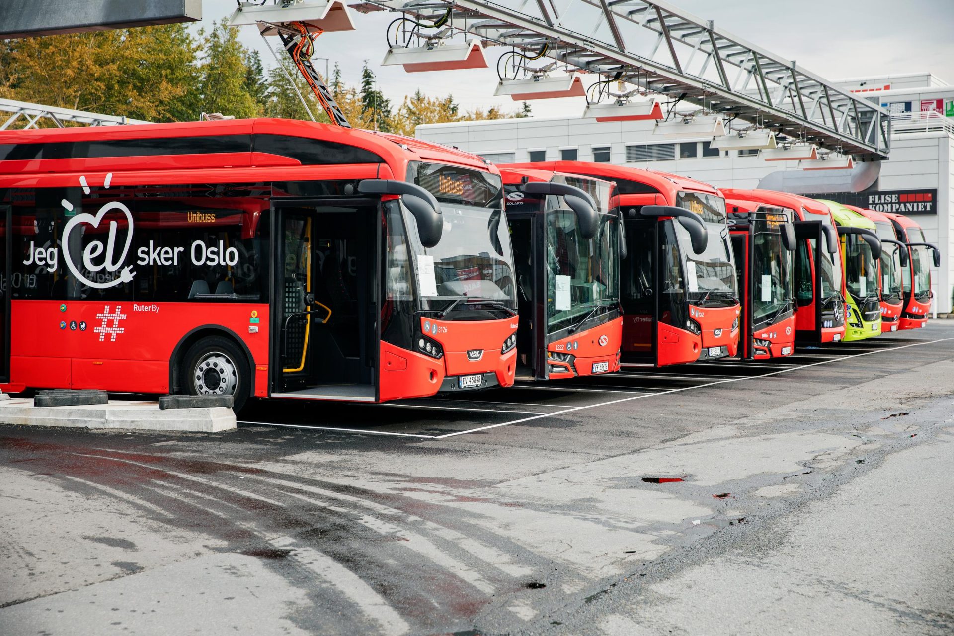 En rad med røde elektriske busser parkert ved ladestasjoner, med slagordet «Jeg elsker oslo» på en av bussene.
