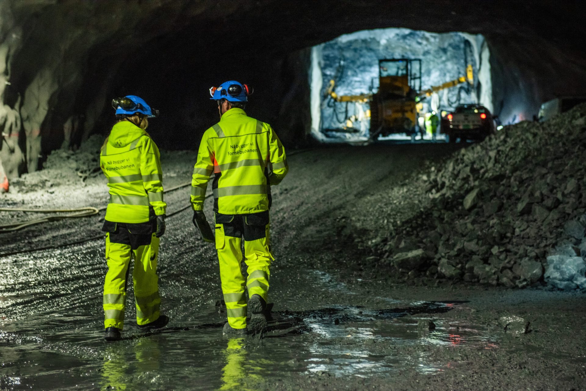 To bygningsarbeidere i gule jakker og bukser går mot tunge maskiner i en svakt opplyst tunnel.