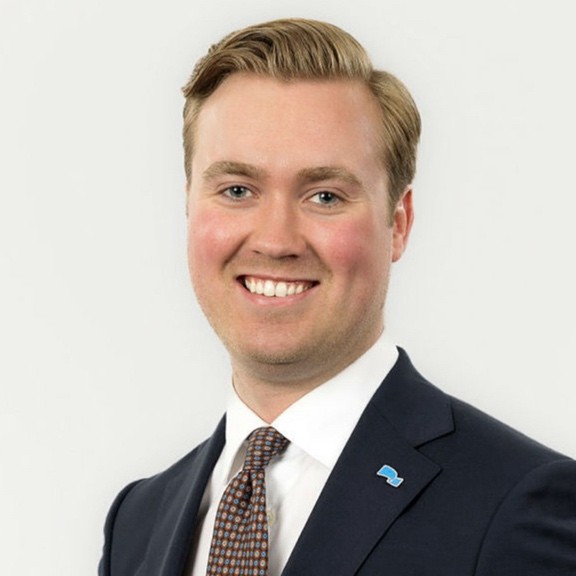 Professional headshot of a smiling man with blond hair, wearing a dark suit, white shirt and patterned tie, with a lapel pin.