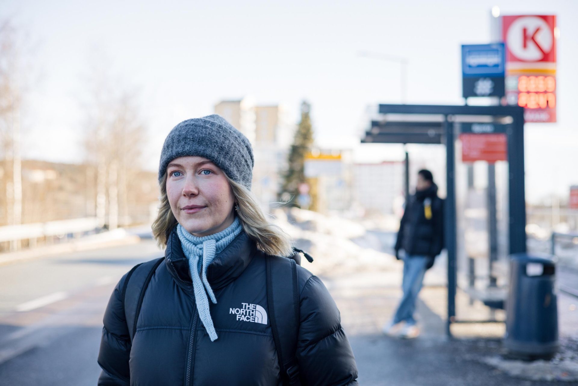 Woman in winter clothes standing at a bus stop with snowy surroundings and blurred background.