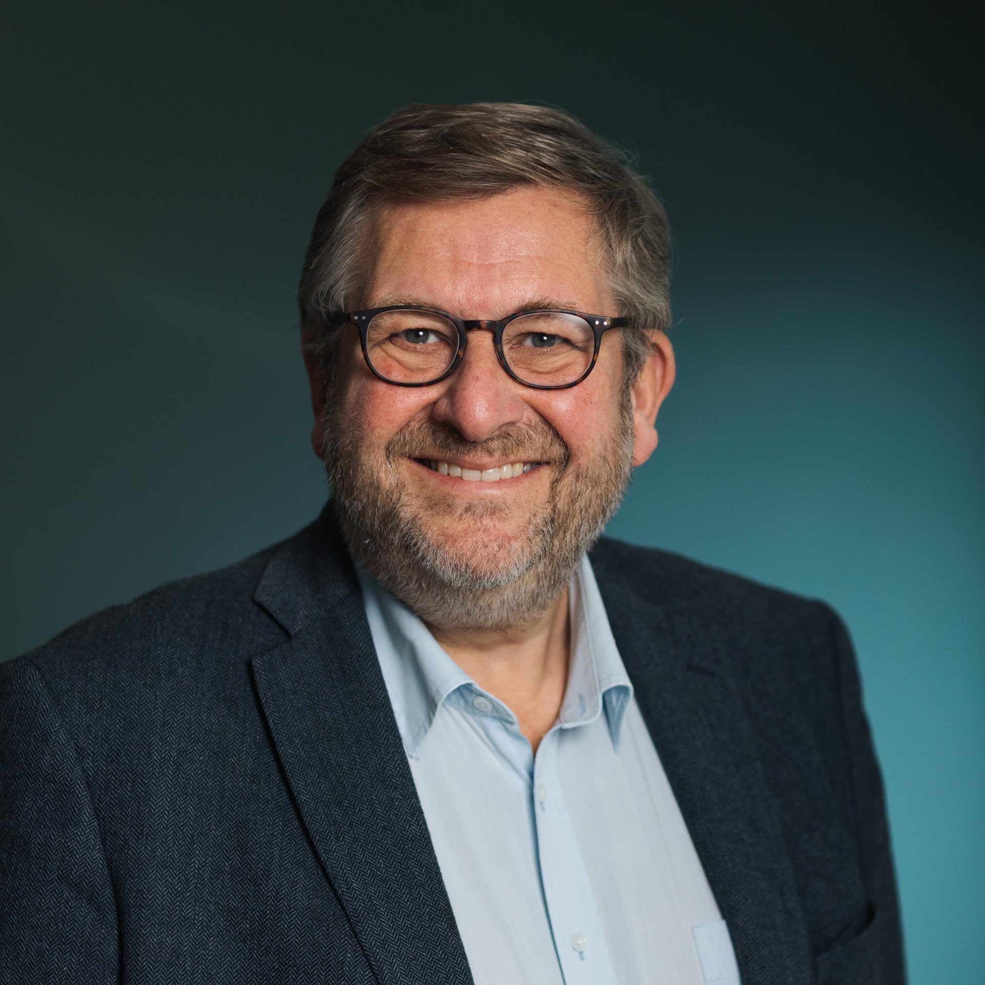 Profesjonelt hodebilde av Ruters administrerende direktør, Bernt Reitan Jenssen, en smilende middelaldrende mann med grått hår og skjegg, iført briller og blå dress over en lyseblå skjorte, mot en blågrønn bakgrunn.