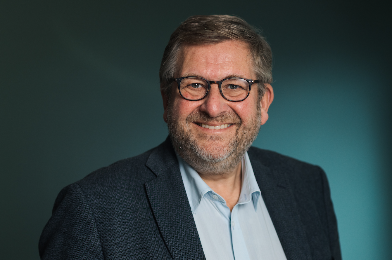 Professional headshot of Ruter's CEO, Bernt Reitan Jenssen, a smiling middle-aged man with gray hair and beard, wearing glasses and a blue suit over a light blue shirt, against a teal background.