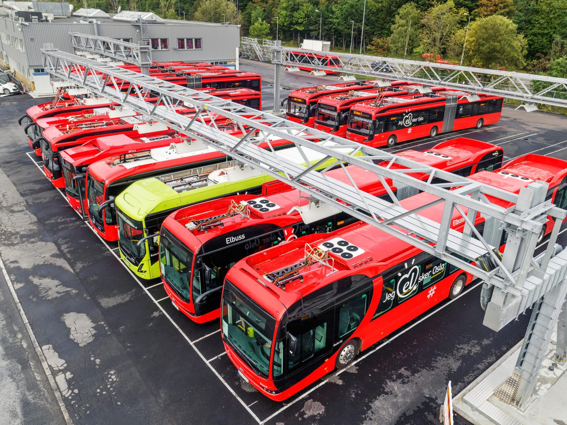 Luftfoto av et bussdepot som viser mange røde og én grønn buss som står til lading.